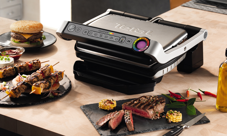 Tefal GC702D Optigrill sw/​geb. edelst. inkl. Hamburgerpresse. auf dem Tisch mit frisch gegrilltem Fleisch.